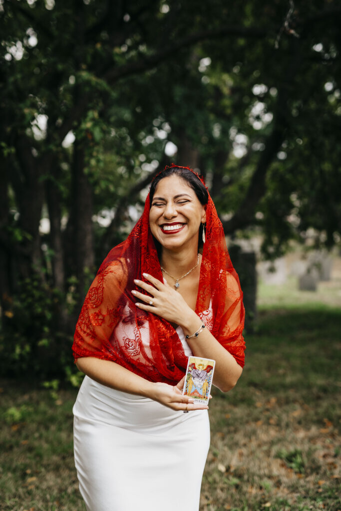 A bride poses in a red veil with the lovers tarot card at a cemetery in Salem Massachusetts